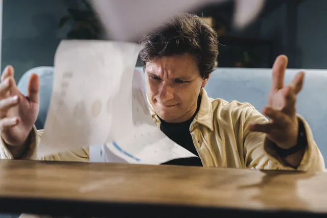 Stressed man throwing documents in frustration at a wooden desk in an indoor setting. Planowanie tygodnia w małej firmie: priorytety, grafik i przegląd zadań
